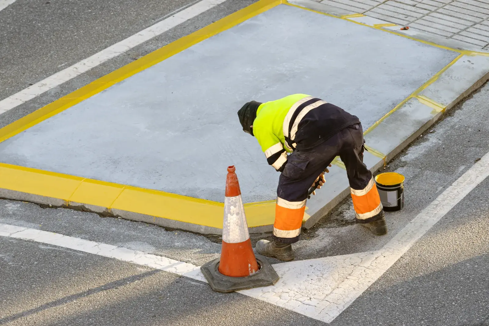 Worker painting a concrete curb edge yellow with a paint bucket and traffic cone beside it